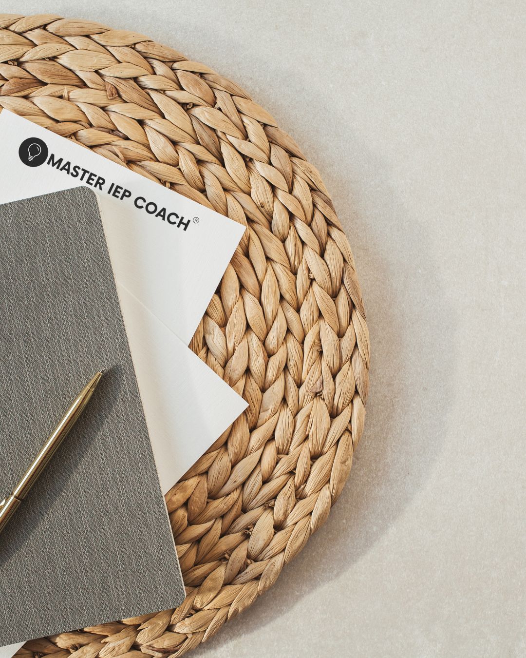 Close-up flat lay of a gold pen resting on a gray textured notebook and a piece of white stationery with the Master IEP Coach® logo. All items are set on a round, woven wicker placemat.